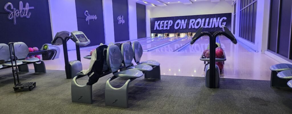 Modern bowling alley interior featuring ergonomic grey seating, digital touch-screen scoring consoles, and a large black and white banner reading 'KEEP ON ROLLING' above the lanes under purple ambient lighting.