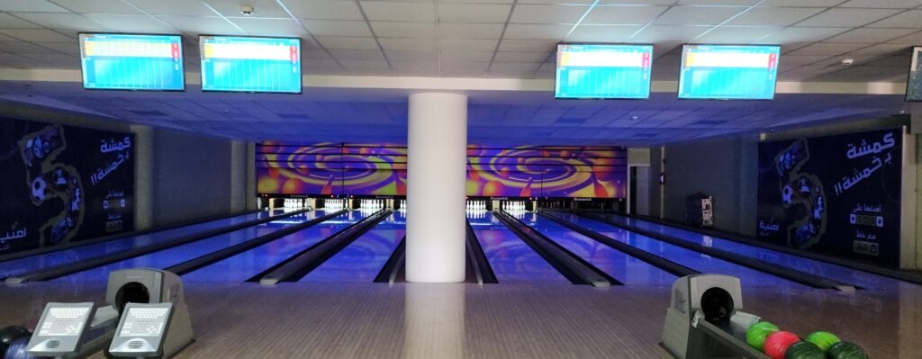A modern bowling alley with 8 lanes illuminated by a bright blue glow. A large white cylindrical pillar stands between the center lanes. In the foreground, ball returns hold red and green bowling balls next to digital consoles. Large black banners with Arabic text and colorful graphics line the side walls, and all pins are set at the end of the polished lanes.