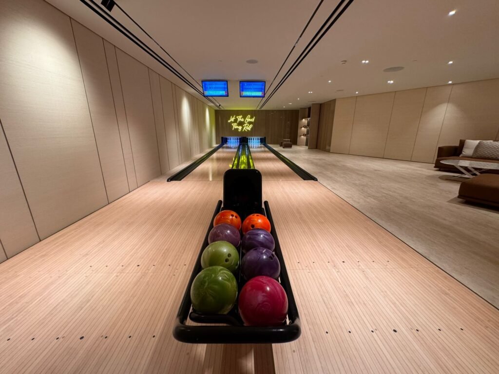 A luxury private two-lane bowling alley featuring minimalist wood paneling, a yellow neon sign reading 'Let The Good Times Roll', and a modern lounge area with a plush sectional sofa.