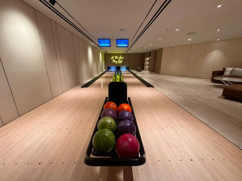 A luxury private two-lane bowling alley featuring minimalist wood paneling, a yellow neon sign reading 'Let The Good Times Roll', and a modern lounge area with a plush sectional sofa.