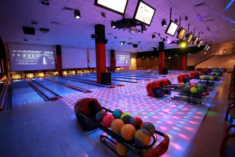 Wide view of a large commercial bowling alley with cosmic neon lighting, pink and blue disco floor patterns, large projector screens at the end of the lanes, and multiple red ball returns with colorful bowling balls.